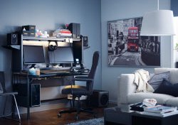 A black workstation with space for two screens, speakers and storage on top, shown inside a living room.