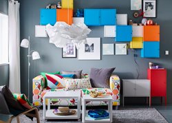 A small living room furnished with a two-seat sofa with a cover in a colorful geometric pattern and two white side tables on castors. Shown together with colorful wall cabinets in different sizes.