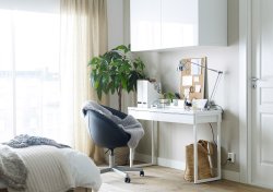 A white desk with a black swivel chair on castors and a wall cabinet, inside the bedroom.
